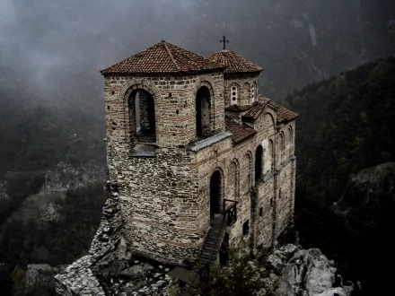 A dark, atmospheric view of Asen's Fortress, featuring the Church of the Holy Mother of God. This beautiful stone structure stands majestically amidst a misty landscape.
