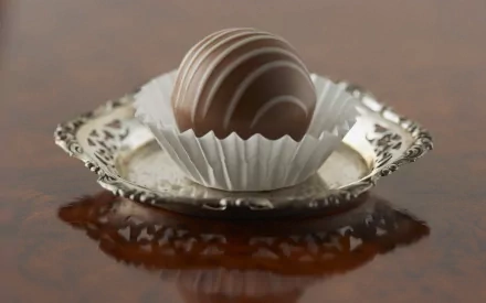 HD desktop wallpaper featuring a close-up of an elegantly swirled chocolate truffle in a white paper cup, sitting on an ornate silver plate against a blurred wooden background.
