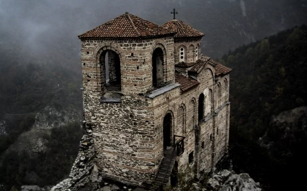 A dark, atmospheric view of Asen's Fortress, featuring the Church of the Holy Mother of God. This beautiful stone structure stands majestically amidst a misty landscape.