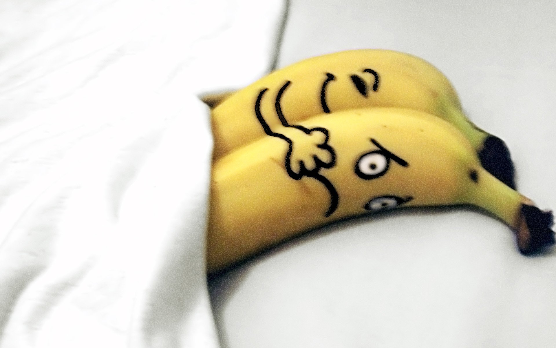 Two bananas with funny faces lying under a blanket, creating a humorous scene. This HD desktop wallpaper adds a playful touch to your background.