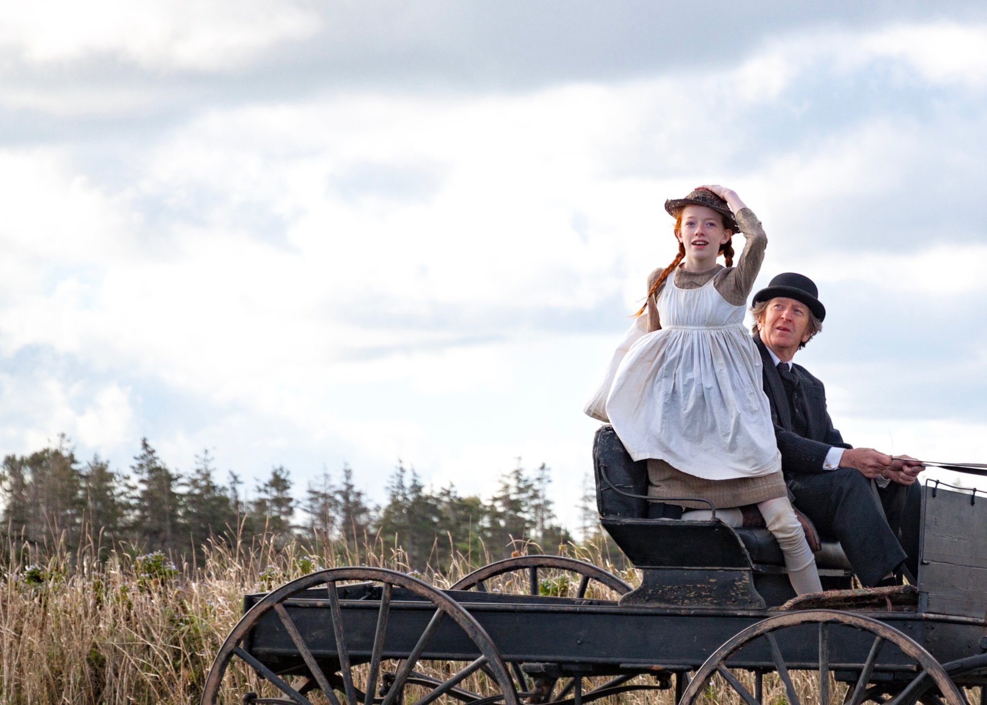HD PC desktop wallpaper from the TV show Anne with an E: red-haired Anne in braids and a pinafore sits in a horse-drawn cart with an older man across a windswept field.