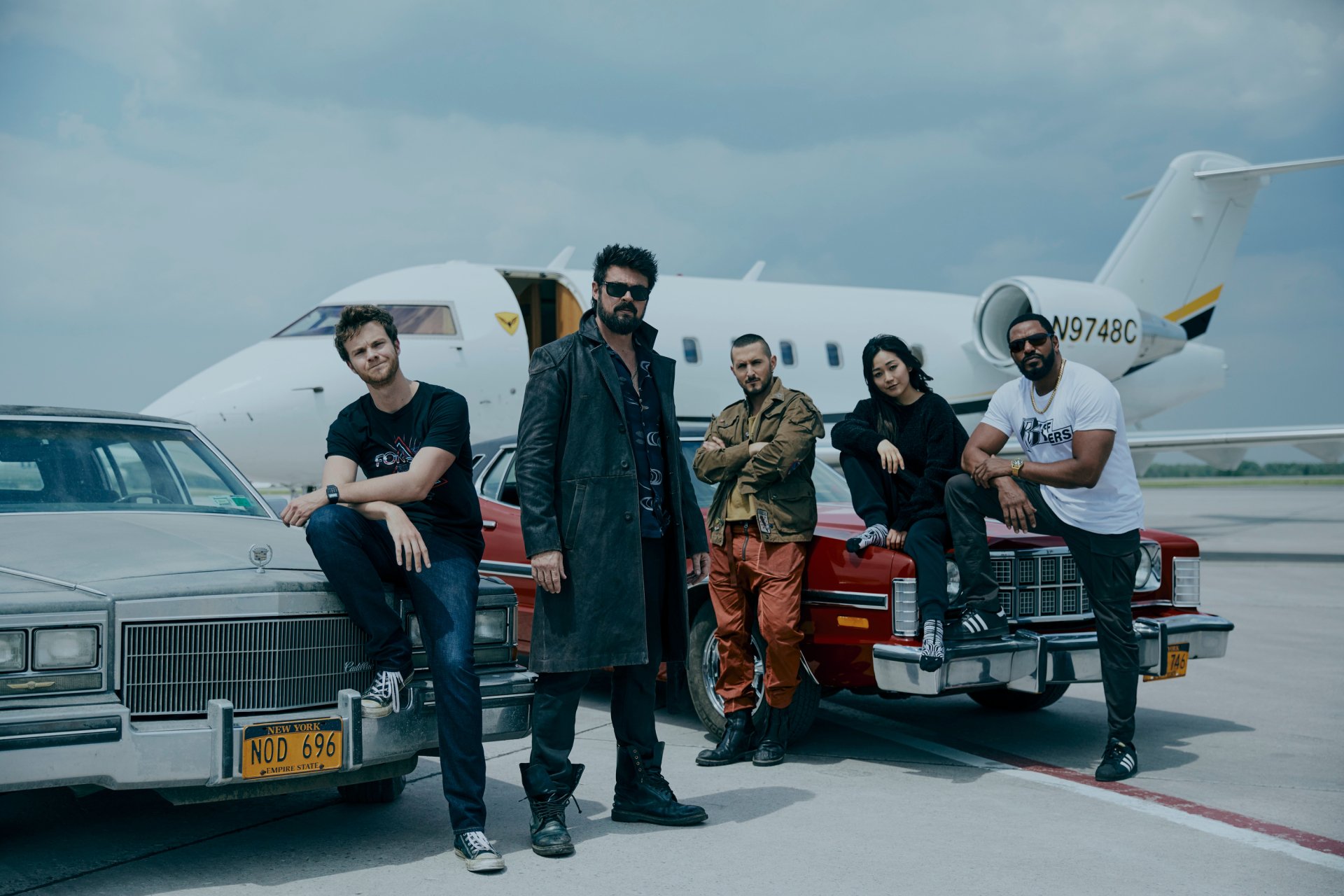 HD PC desktop wallpaper featuring the main cast of the TV show The Boys (2019) standing and sitting near cars on an airport runway under a cloudy sky.