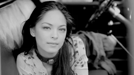 Black and white HD desktop wallpaper featuring celebrity Kristin Kreuk with a calm expression, seated in a softly lit environment.