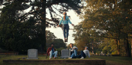Stranger Things TV scene 2K Quad HD PC wallpaper: Eleven levitates above a cemetery while friends sit on the grass among tombstones beneath tall trees.
