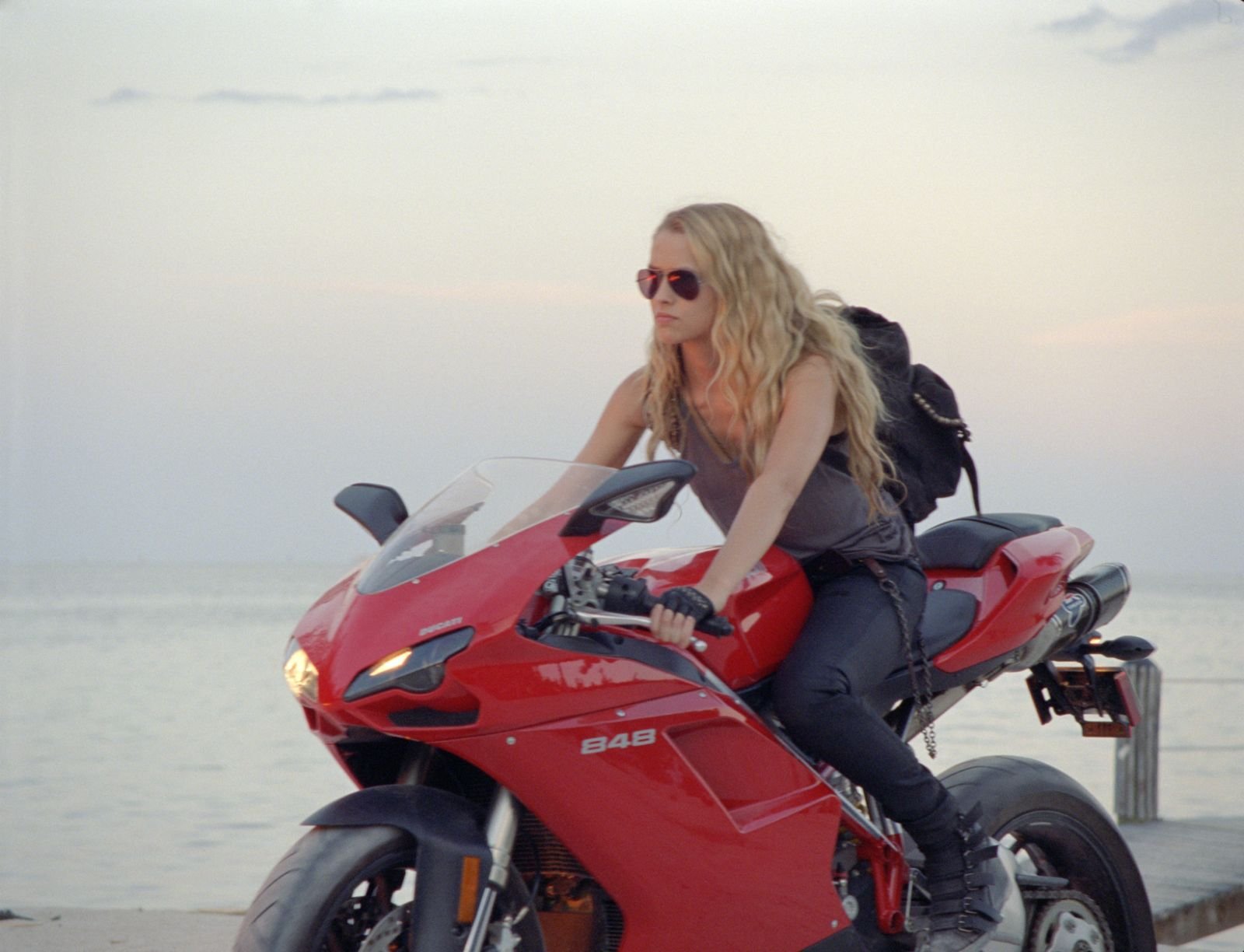 A woman with long hair and sunglasses sits confidently on a red motorcycle against a serene ocean background, inspired by the movie I Am Number Four.