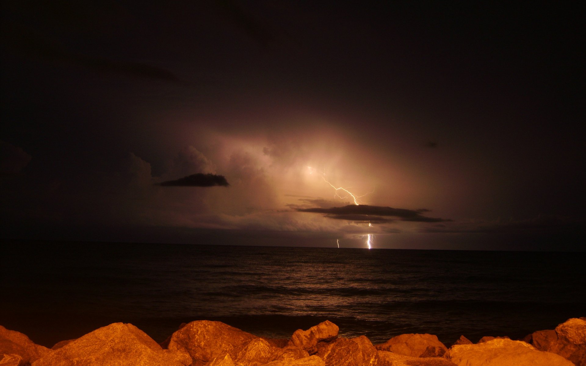 HD desktop wallpaper of a night ocean scene with lightning illuminating distant clouds, casting a dramatic light over calm waters and rocky shoreline.