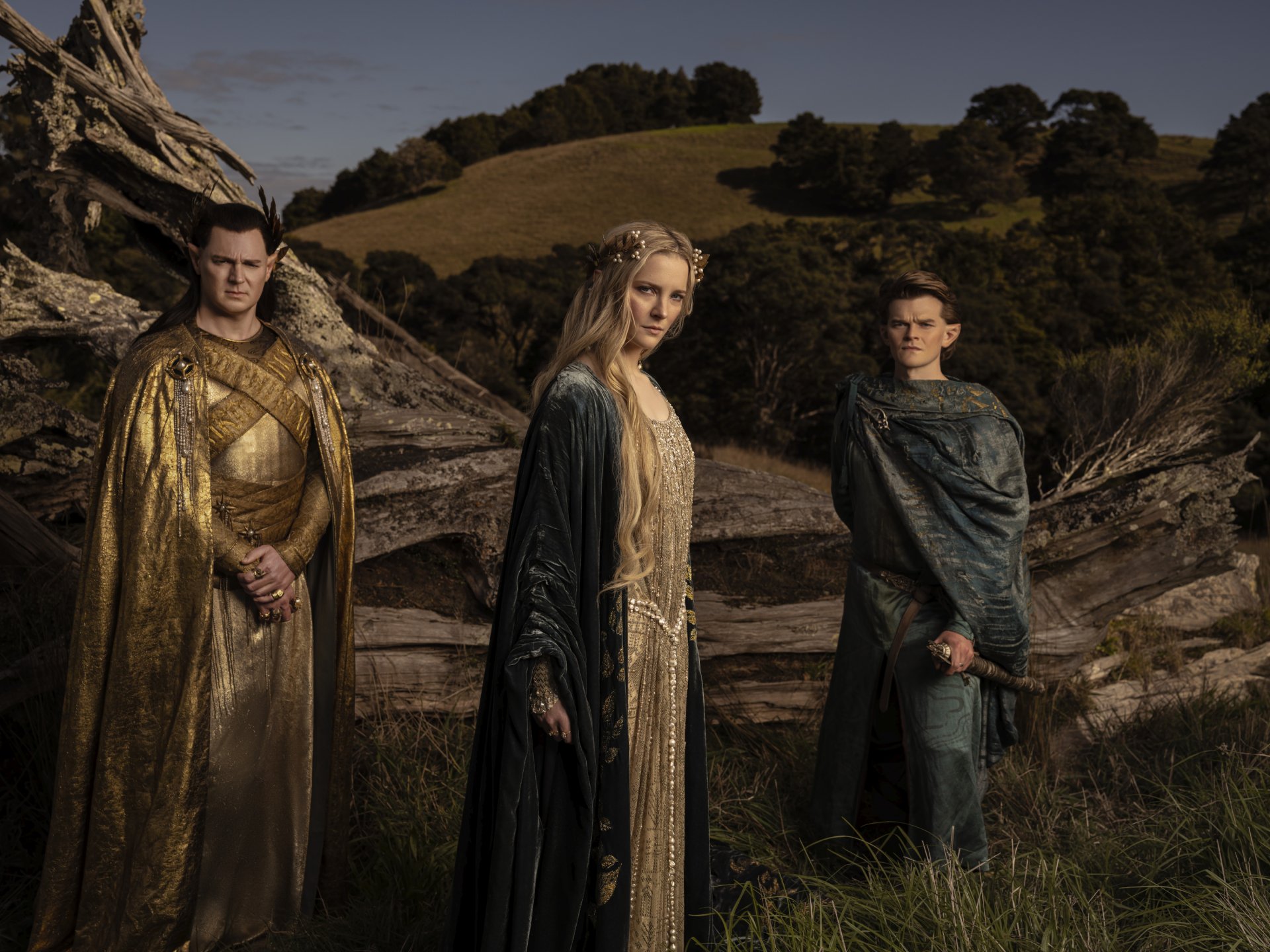 2K Quad HD PC desktop wallpaper: three robed characters from the TV show The Lord of the Rings: The Rings of Power stand in a windswept grassy landscape, clad in gold and green ceremonial robes.