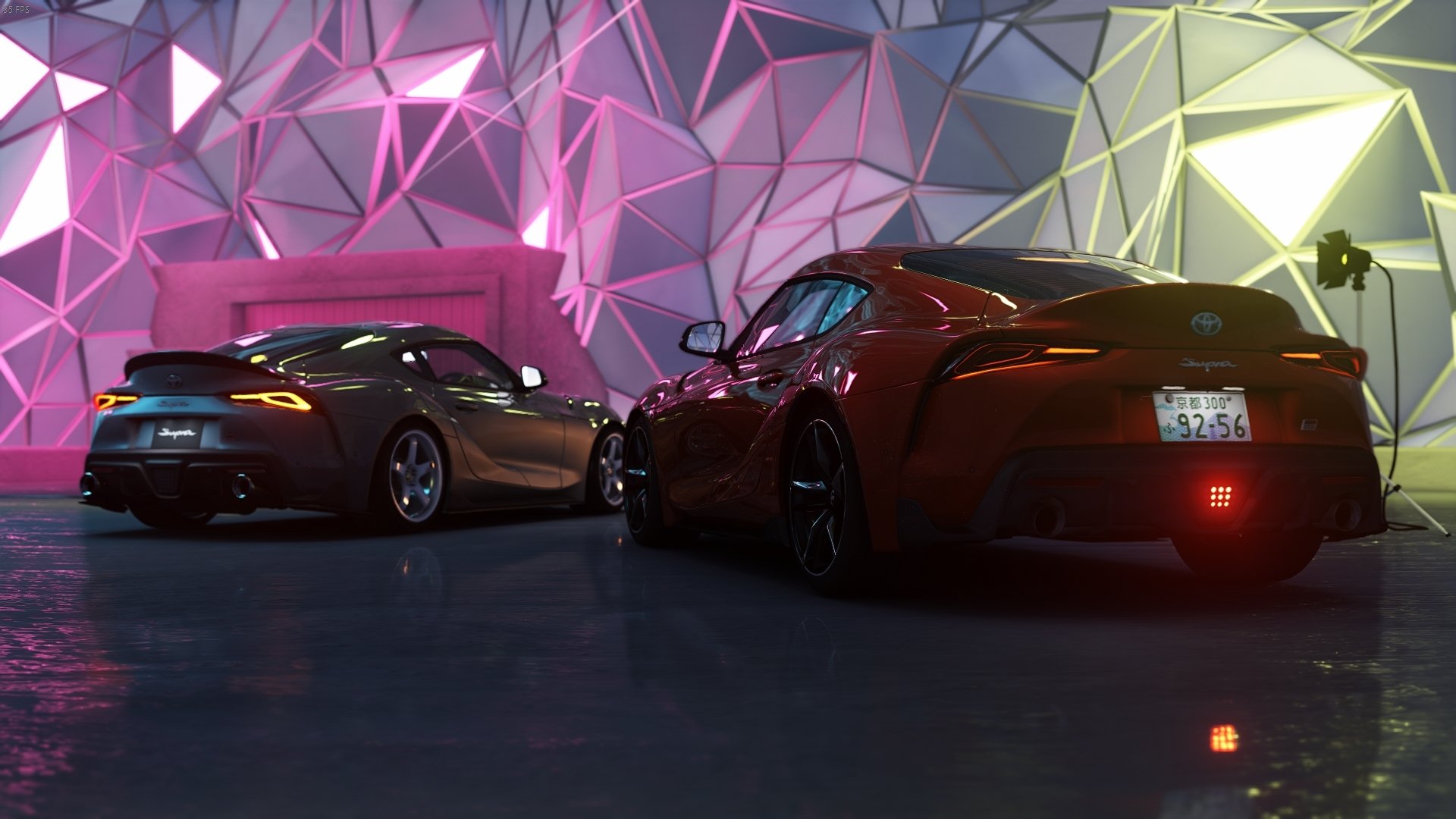HD PC desktop wallpaper of two Toyota Supras in a neon-lit showroom from Assetto Corsa, rear three-quarter view with angular pink and green illuminated geometric wall panels.
