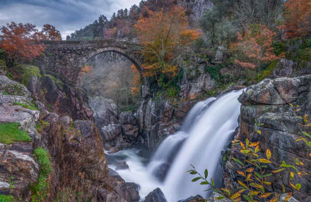 A 4K Ultra HD image of a man-made stone bridge over a waterfall surrounded by autumn foliage in Portugal.