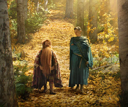 HD PC desktop wallpaper from the TV show The Lord of the Rings: The Rings of Power — two robed figures, one short with a long beard, walking a sunlit golden‑leaf forest path.