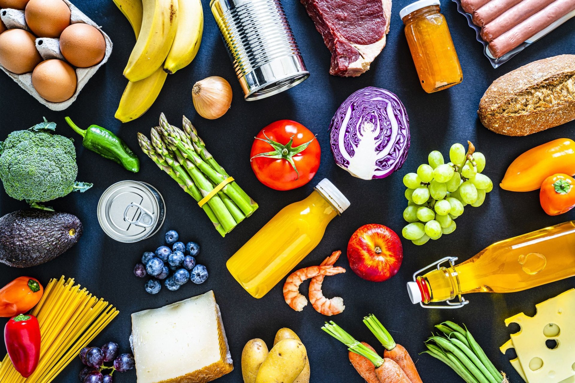 HD PC desktop wallpaper showing a food still life: colorful fruits, vegetables, bread, cheese, eggs, canned goods and bottles arranged on a dark background.