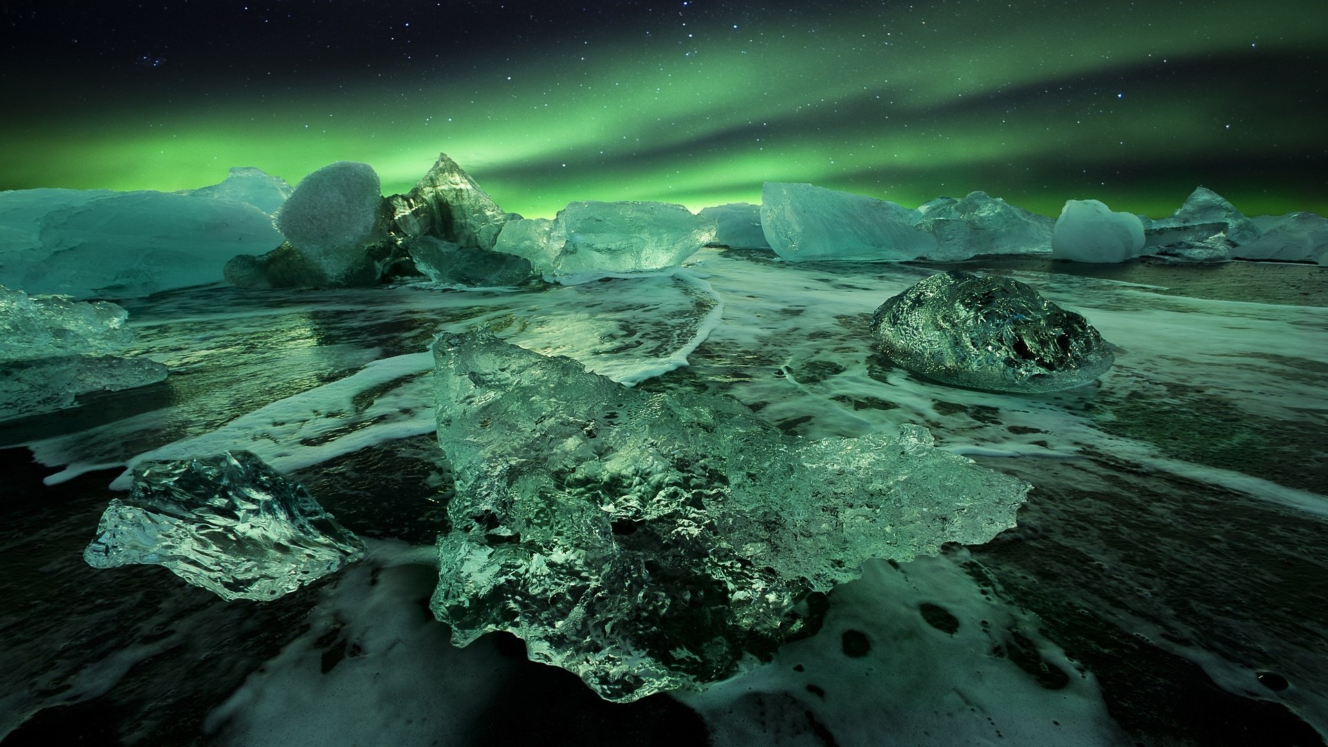 HD PC desktop wallpaper/background: nature scene of glassy ice chunks on a dark shore beneath a vivid green aurora borealis.