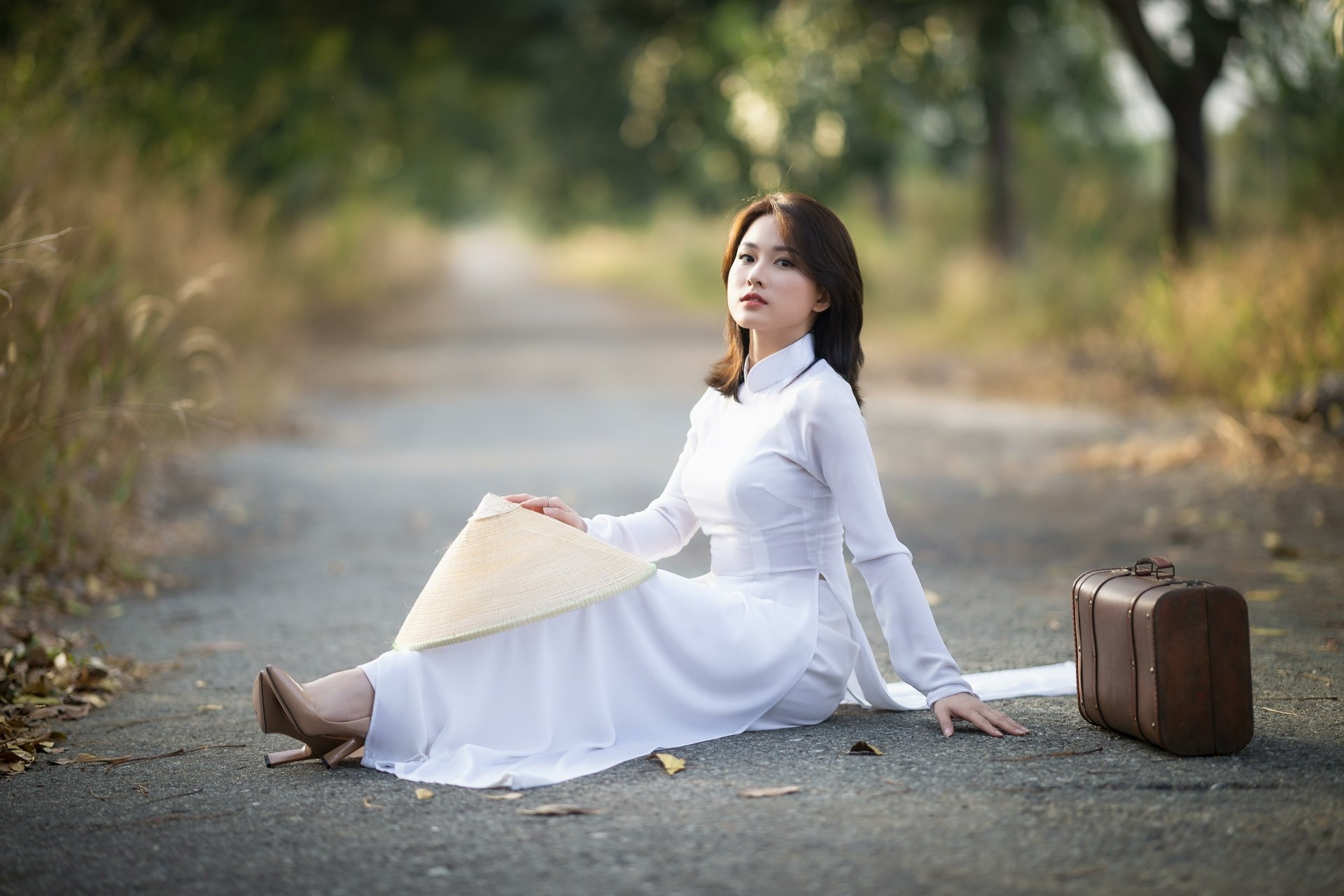 4K Ultra HD PC desktop wallpaper: a woman in a white flowing dress seated on a tree-lined country road, holding a conical hat with a vintage suitcase at her side.