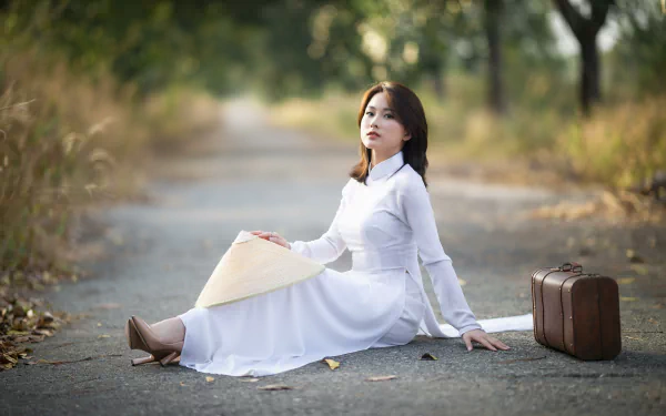 4K Ultra HD PC desktop wallpaper: a woman in a white flowing dress seated on a tree-lined country road, holding a conical hat with a vintage suitcase at her side.