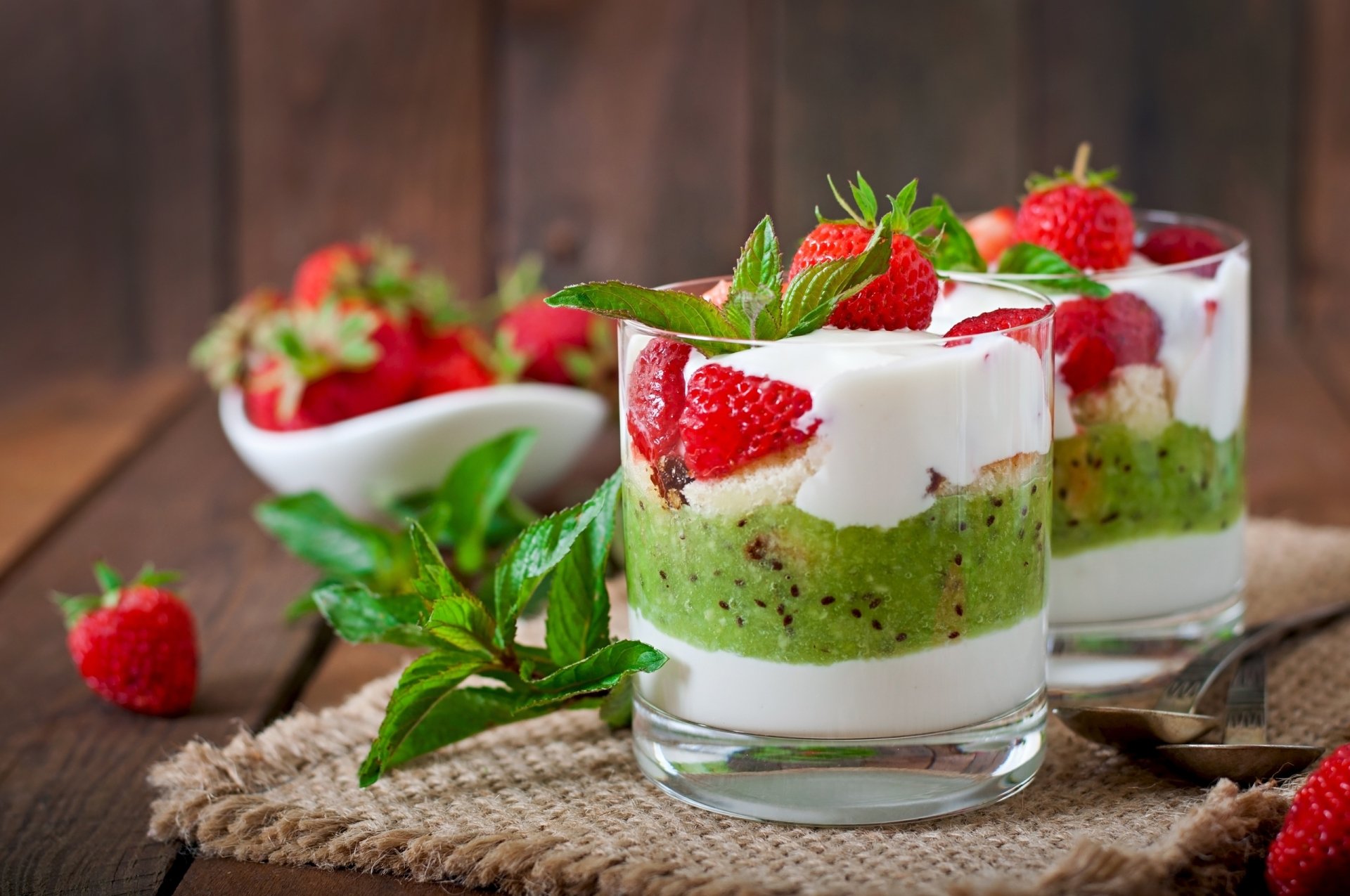 HD desktop wallpaper featuring two glasses of layered dessert with green sponge cake, white cream, and fresh strawberries topped with mint leaves.