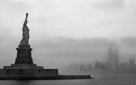 A black and white HD image of the Statue of Liberty standing majestically on its pedestal, shrouded in mist with the New York City skyline faintly visible in the background.