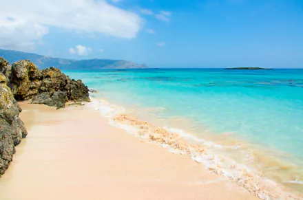 HD desktop wallpaper image featuring a serene beach with golden sand, azure water, and rocky shore under a clear blue sky. The background showcases distant mountains and tranquil nature.