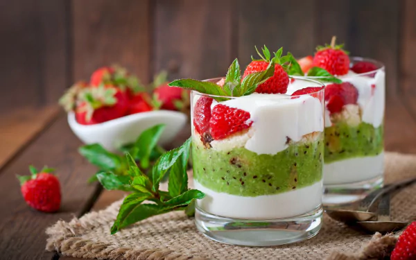 HD desktop wallpaper featuring two glasses of layered dessert with green sponge cake, white cream, and fresh strawberries topped with mint leaves.