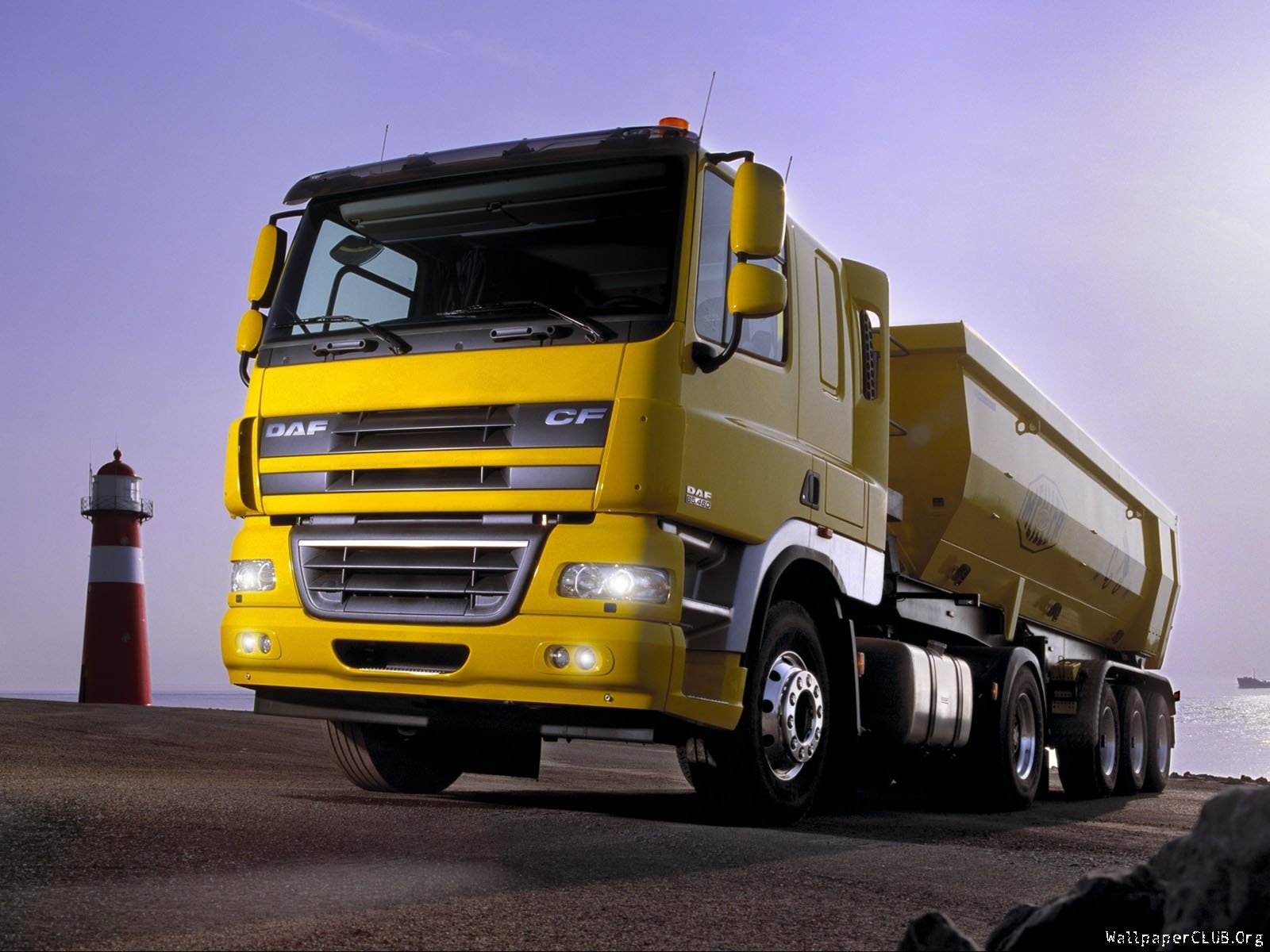 HD PC desktop wallpaper/background showing a yellow DAF heavy-duty truck (vehicle) parked by a lighthouse under a dusky purple sky.
