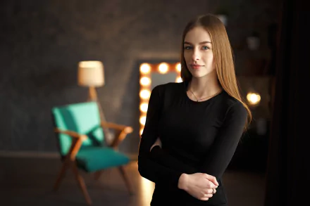 Young woman model in a black top with arms crossed, posed in a dim studio with a lit mirror and teal chair — 2K Quad HD PC desktop wallpaper/background.