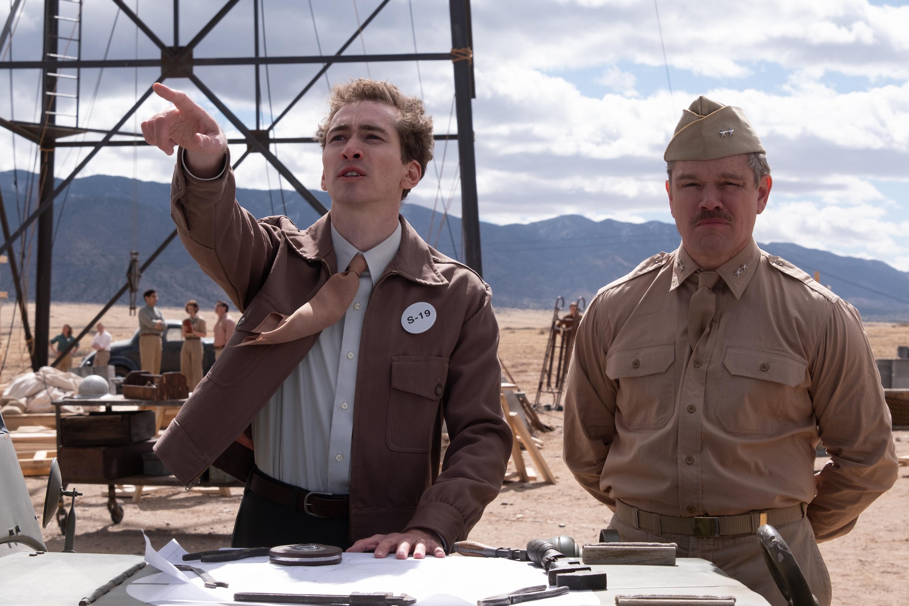 HD wallpaper featuring actors in character on the set of the movie Oppenheimer, with one gesturing upwards and the other standing beside him in a military uniform, set against a desert backdrop.