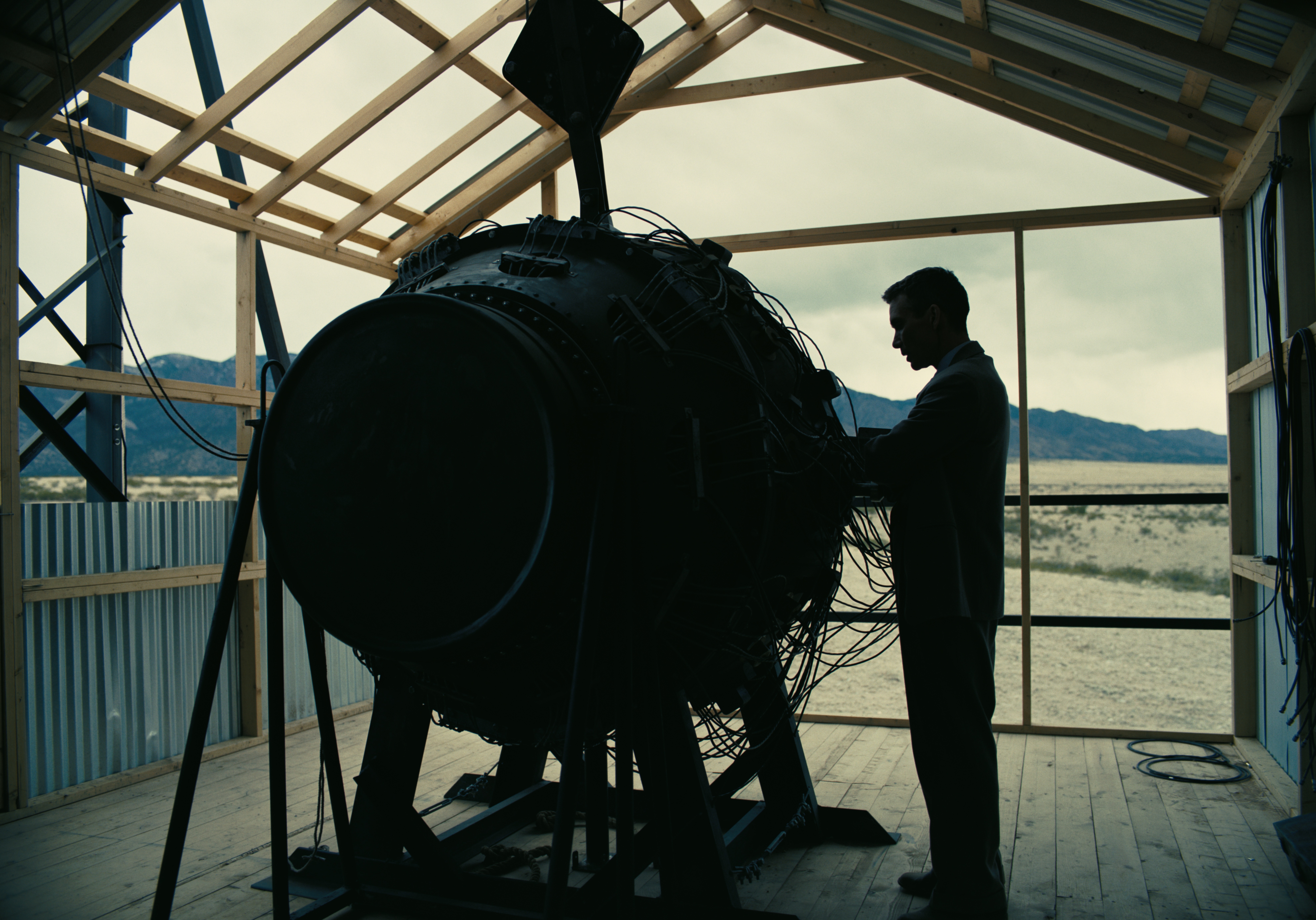 Silhouette of a man resembling an actor, standing next to a large scientific apparatus in a wooden structure, with mountains in the background, as a cinematic HD desktop wallpaper.