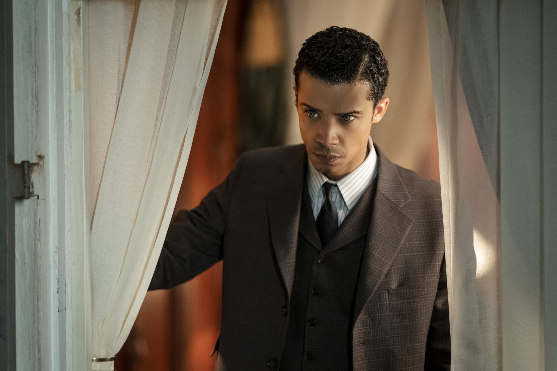 HD desktop wallpaper featuring a character from Interview with the Vampire, portrayed by actor Jacob Anderson, elegantly dressed and peering through curtains with an intense gaze.