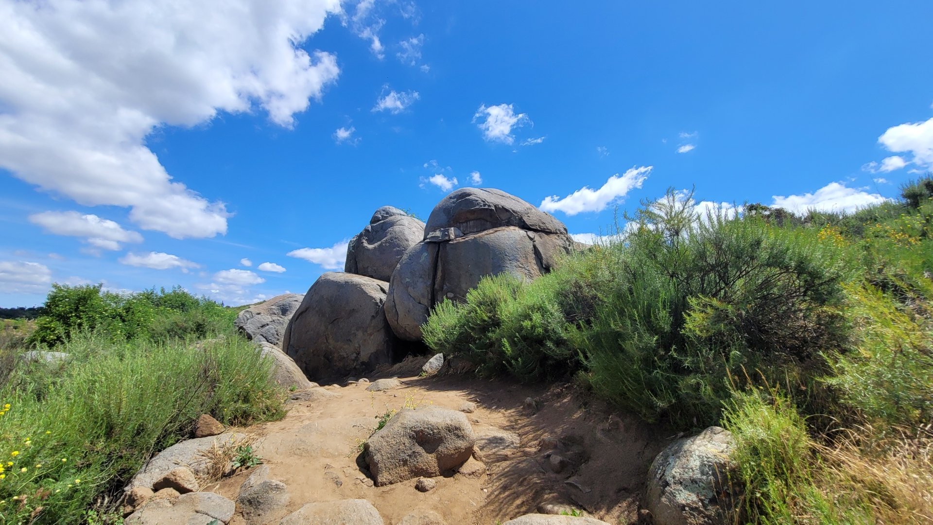 4K Ultra HD landscape of a Southern California hiking trail with large boulders, green shrubs, and a bright blue sky with scattered clouds.