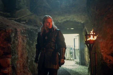 HD desktop wallpaper featuring a character from The Witcher series standing in a dimly lit medieval corridor, next to a lit torch on the wall.