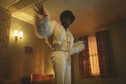 HD desktop wallpaper featuring a character portrayed by Paapa Essiedu in a scene from Black Mirror, dressed in a stylish white outfit with a fur coat, gesturing dramatically in a vintage room setting.
