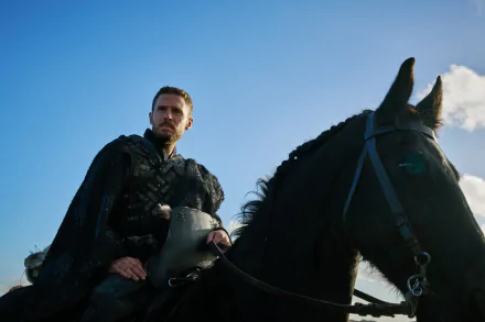 Iain De Caestecker as a character from The Winter King, sits on a black horse against a blue sky, exuding a powerful presence in this HD desktop wallpaper.