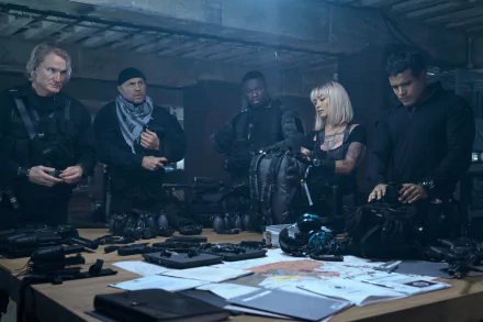 HD wallpaper featuring a group of actors from The Expendables movie series gathered around a table filled with weaponry and tactical gear, portraying a moment of intense planning or debriefing.