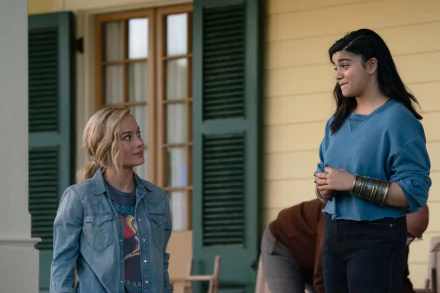 HD desktop wallpaper featuring two characters from The Marvels, with one wearing a denim jacket and the other in a blue sweater, standing in front of a house with green shutters.