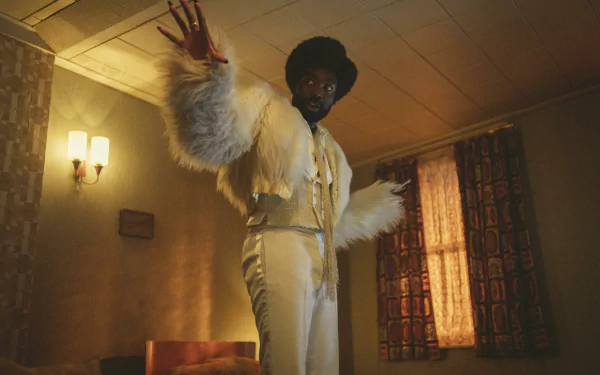 HD desktop wallpaper featuring a character portrayed by Paapa Essiedu in a scene from Black Mirror, dressed in a stylish white outfit with a fur coat, gesturing dramatically in a vintage room setting.