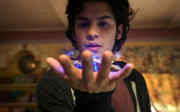 HD wallpaper featuring a young man portraying Jaime Reyes as Blue Beetle, focusing intently on a glowing blue energy emanating from his hand, with a colorful backdrop adding depth and intrigue to the scene.