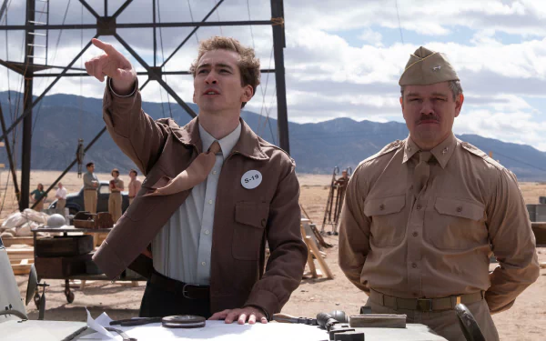 HD wallpaper featuring actors in character on the set of the movie Oppenheimer, with one gesturing upwards and the other standing beside him in a military uniform, set against a desert backdrop.