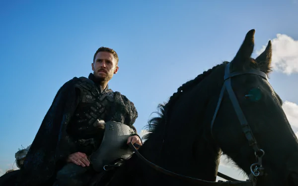 Iain De Caestecker as a character from The Winter King, sits on a black horse against a blue sky, exuding a powerful presence in this HD desktop wallpaper.