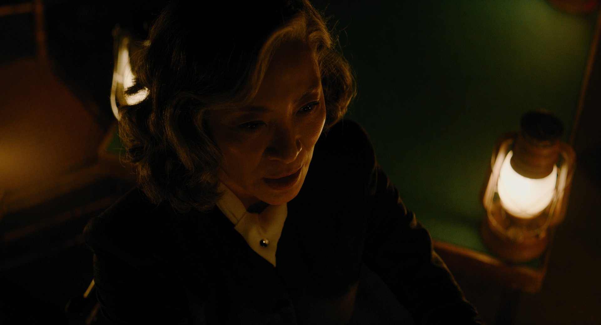 A haunting moment from the movie A Haunting in Venice, featuring a woman illuminated by warm lantern light, creating an eerie and suspenseful atmosphere. HD wallpaper for movie fans.