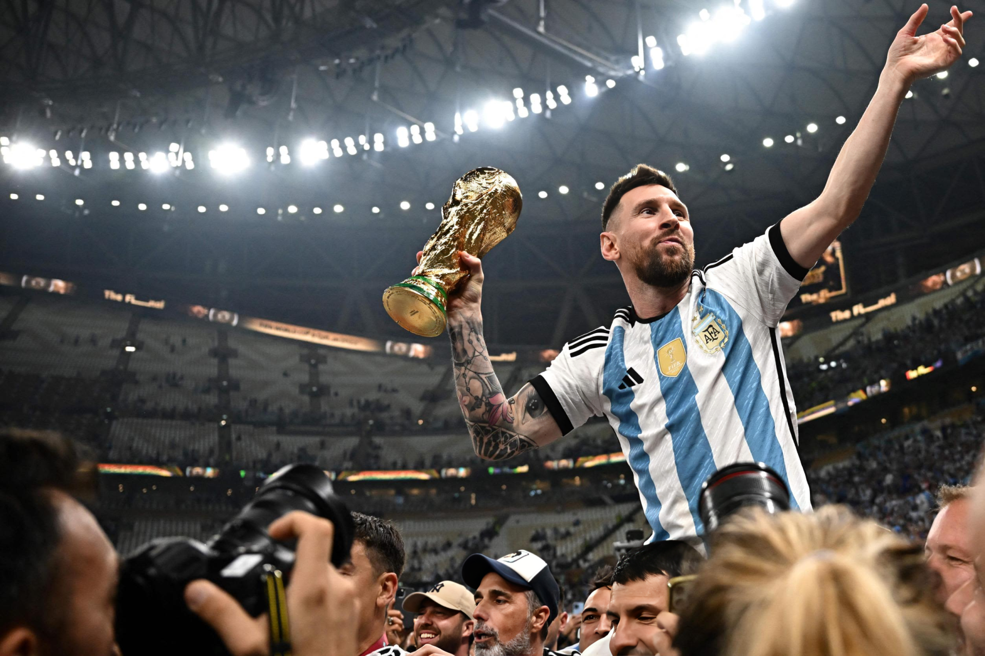 Lionel Messi in Argentina national football team jersey, triumphantly holding a trophy aloft, surrounded by cheering fans in a stadium, captured as HD desktop wallpaper.