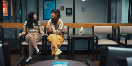 HD wallpaper featuring two women seated and conversing in a waiting room with a Get the shot poster on the wall, tagged as Quiz Lady for desktop backgrounds.