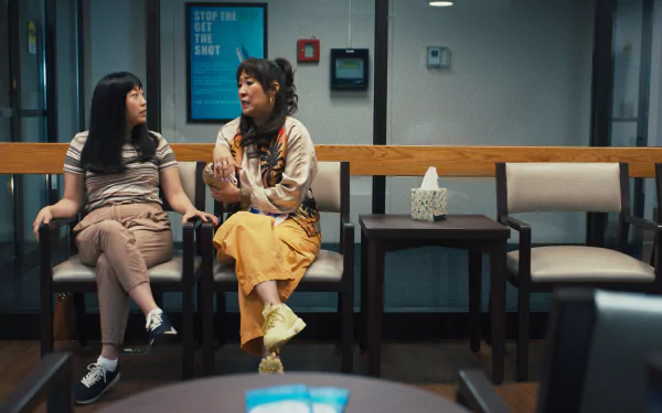 HD wallpaper featuring two women seated and conversing in a waiting room with a Get the shot poster on the wall, tagged as Quiz Lady for desktop backgrounds.
