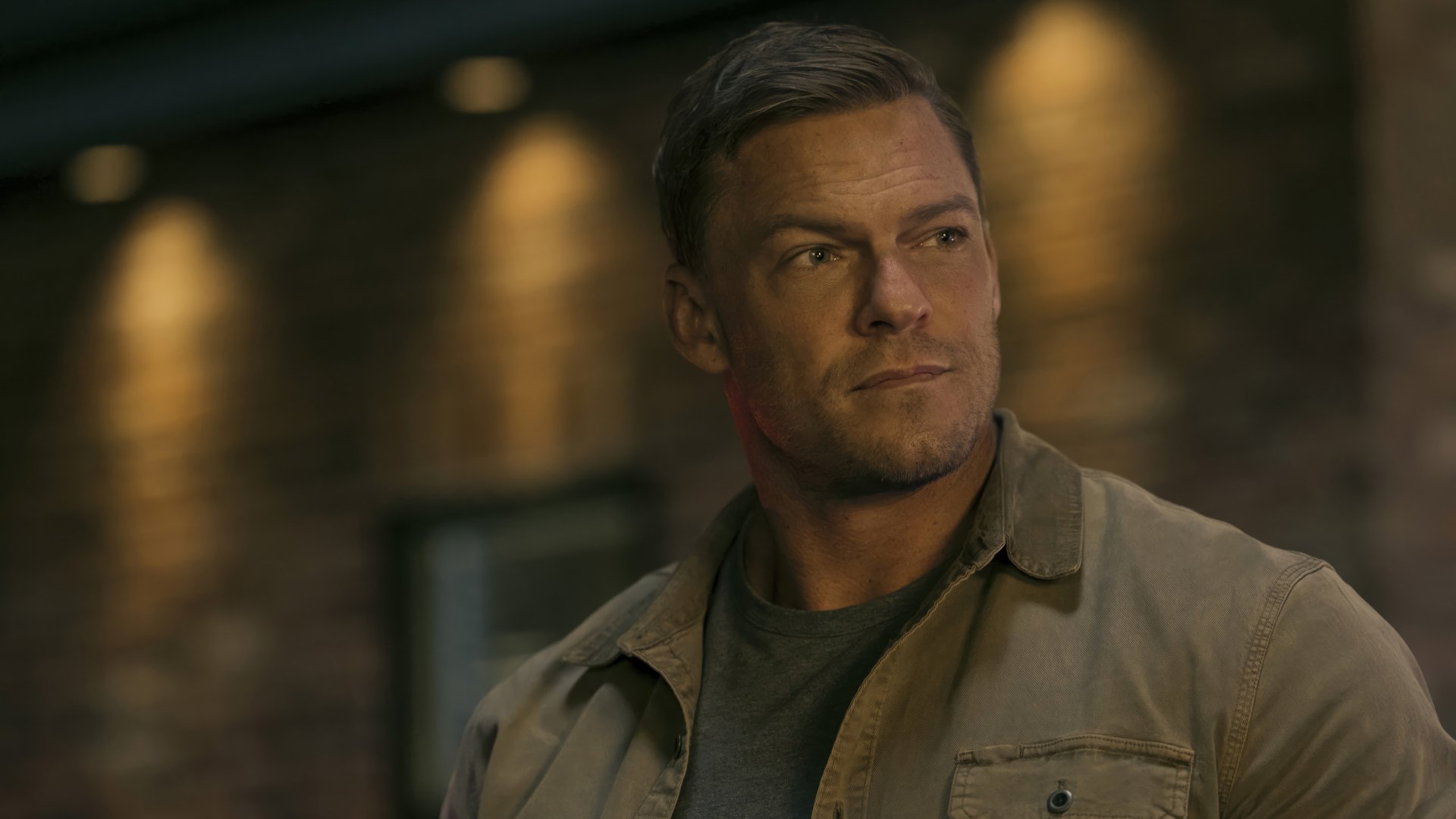 HD wallpaper of Alan Ritchson from the TV show Reacher, posing thoughtfully in a beige jacket against a dark interior background.
