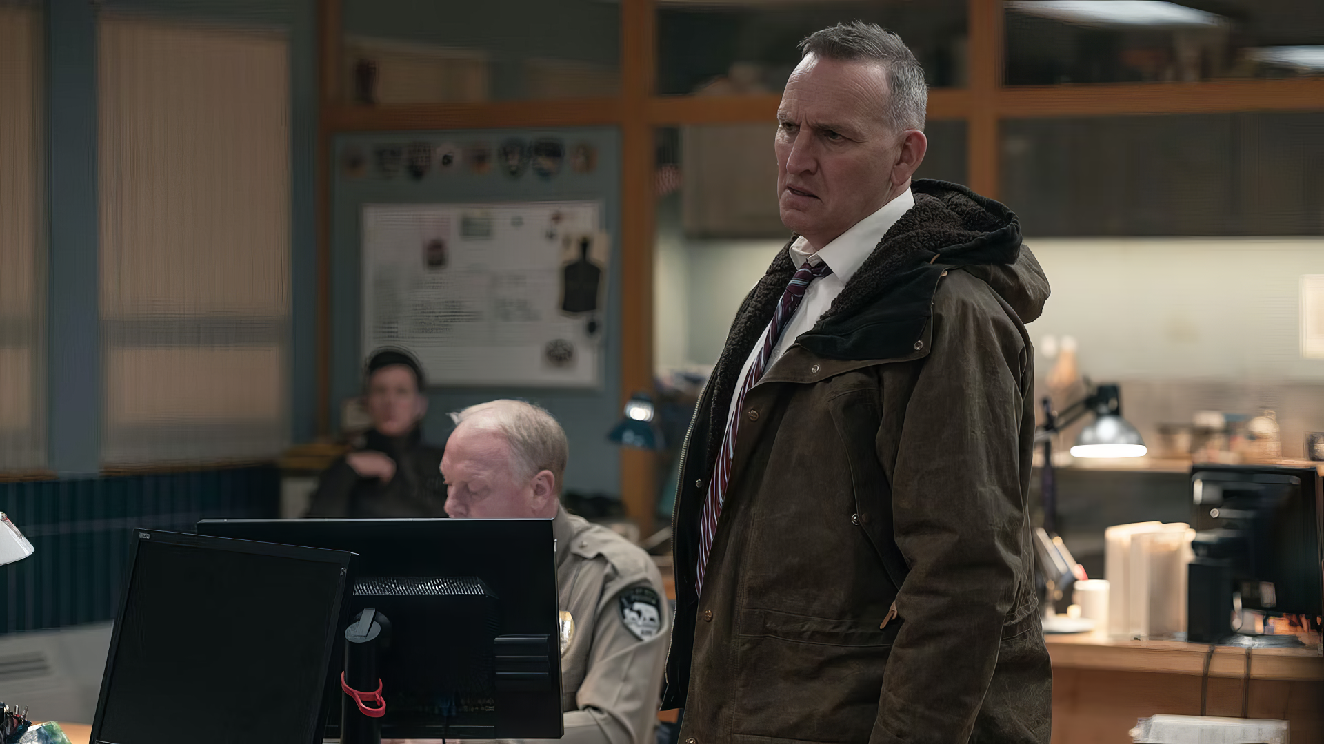 A scene from the TV show True Detective, featuring a serious character in a brown jacket standing in a dimly lit office, with two others engaged in discussion at a computer. Ideal HD desktop wallpaper.