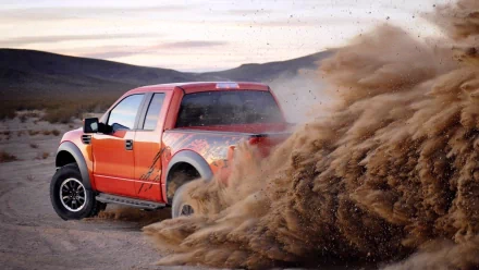 Red Ford Raptor kicking up dirt on a rugged desert trail, captured in a high-definition PC desktop wallpaper.