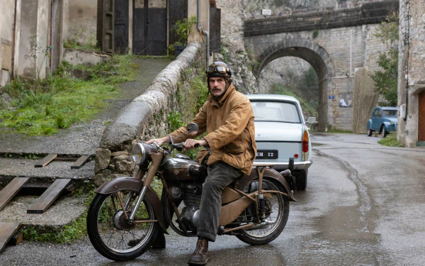 A character from the TV show Monsieur Spade rides a vintage motorcycle on a cobblestone street, with an old stone archway in the background, in a high-definition desktop wallpaper.