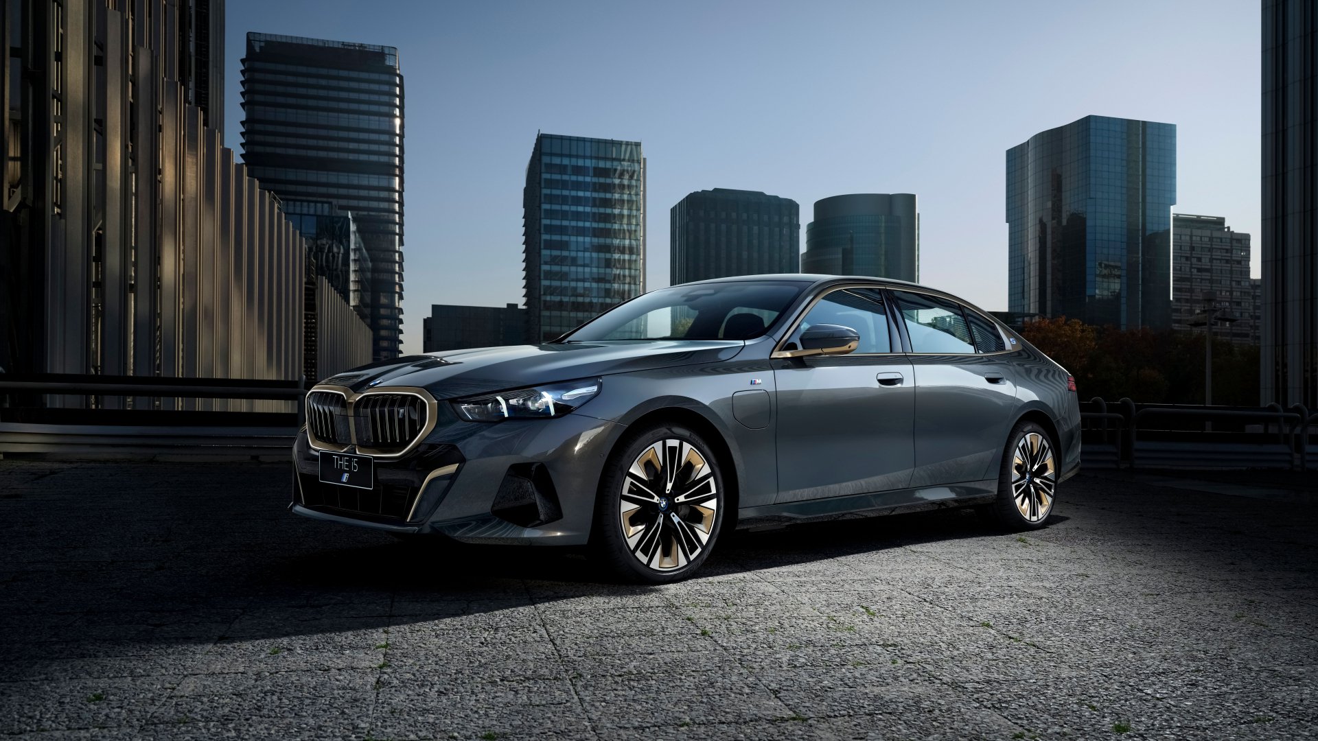 HD desktop wallpaper featuring a BMW i5 M Sport parked in an urban setting with skyscrapers in the background, highlighting the vehicle's sleek design and modern aesthetics.