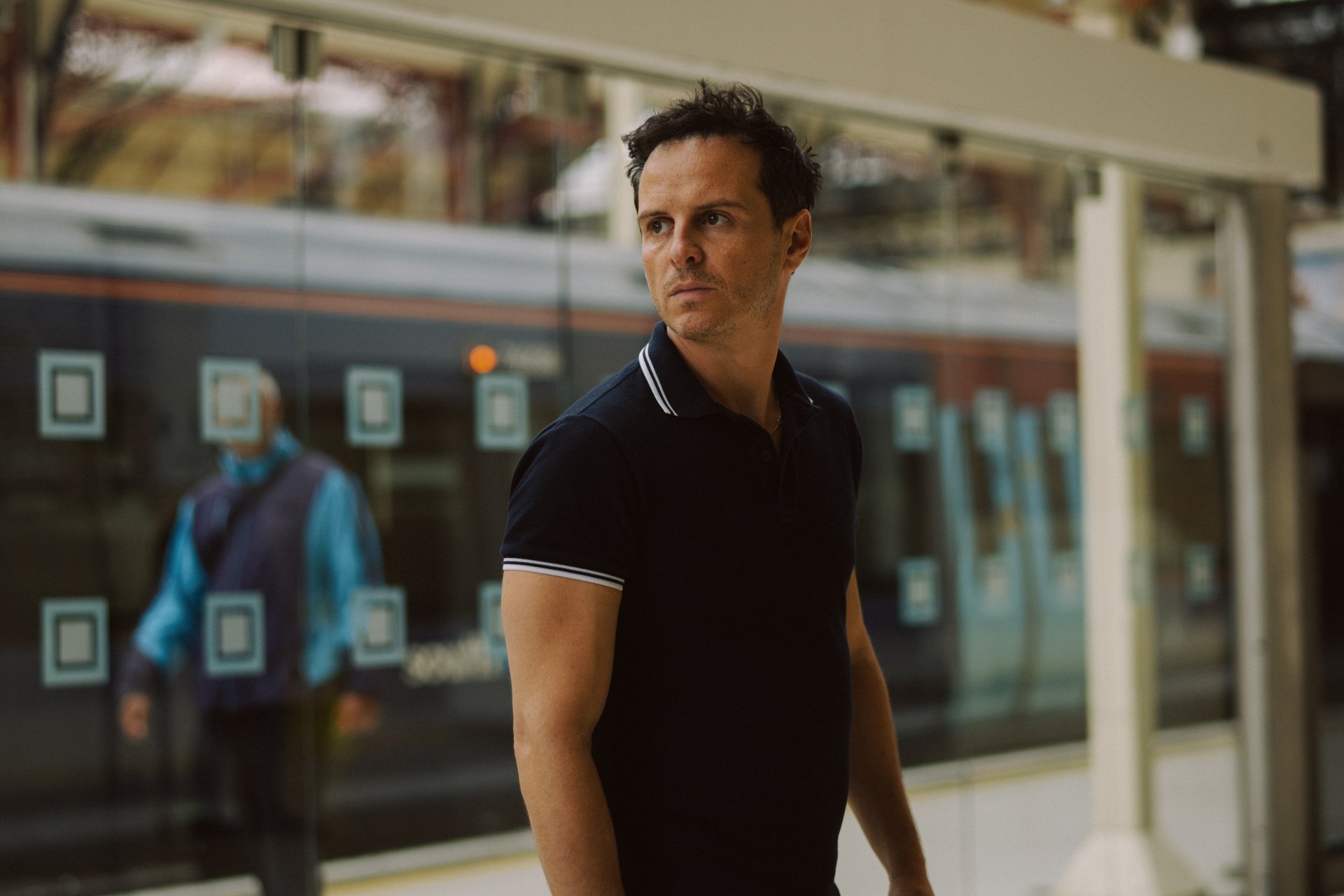 A scene from the movie *All of Us Strangers*, featuring a man in a dark polo shirt, gazing thoughtfully while standing near a train station backdrop. HD desktop wallpaper and background.