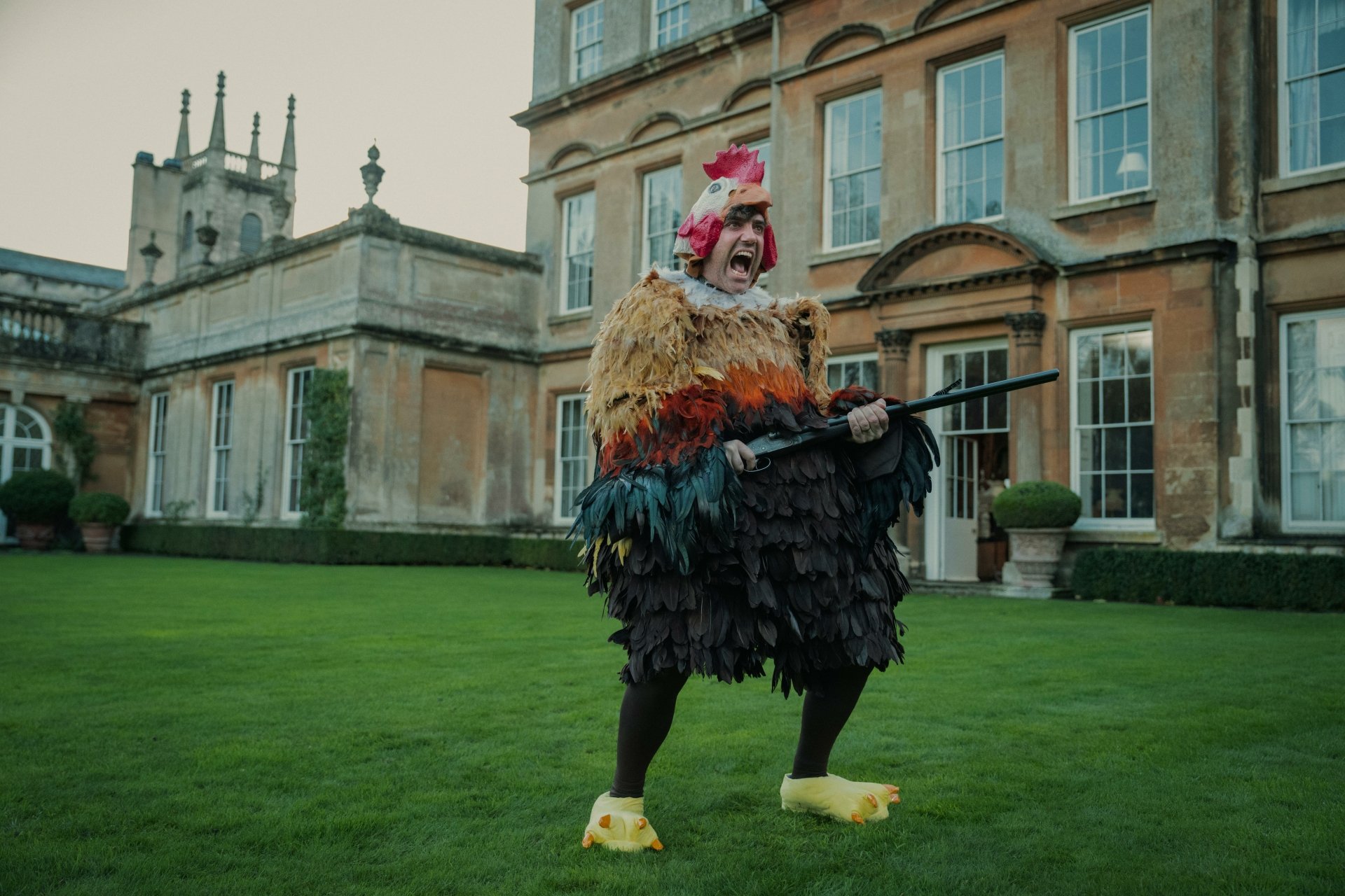Quirky costume from The Gentlemen (2024) TV show showcased in an HD desktop wallpaper featuring a character dressed as a colorful bird in front of an elegant manor house.