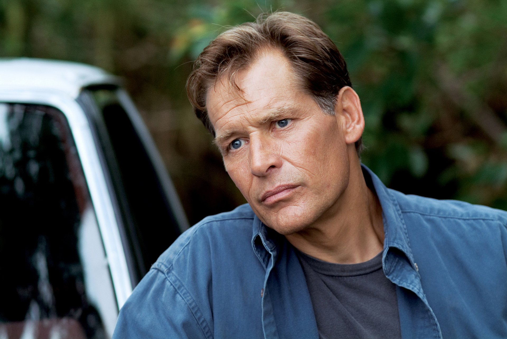 HD desktop wallpaper of a character from the TV show Dexter with a contemplative expression, standing by a car in a forested area.