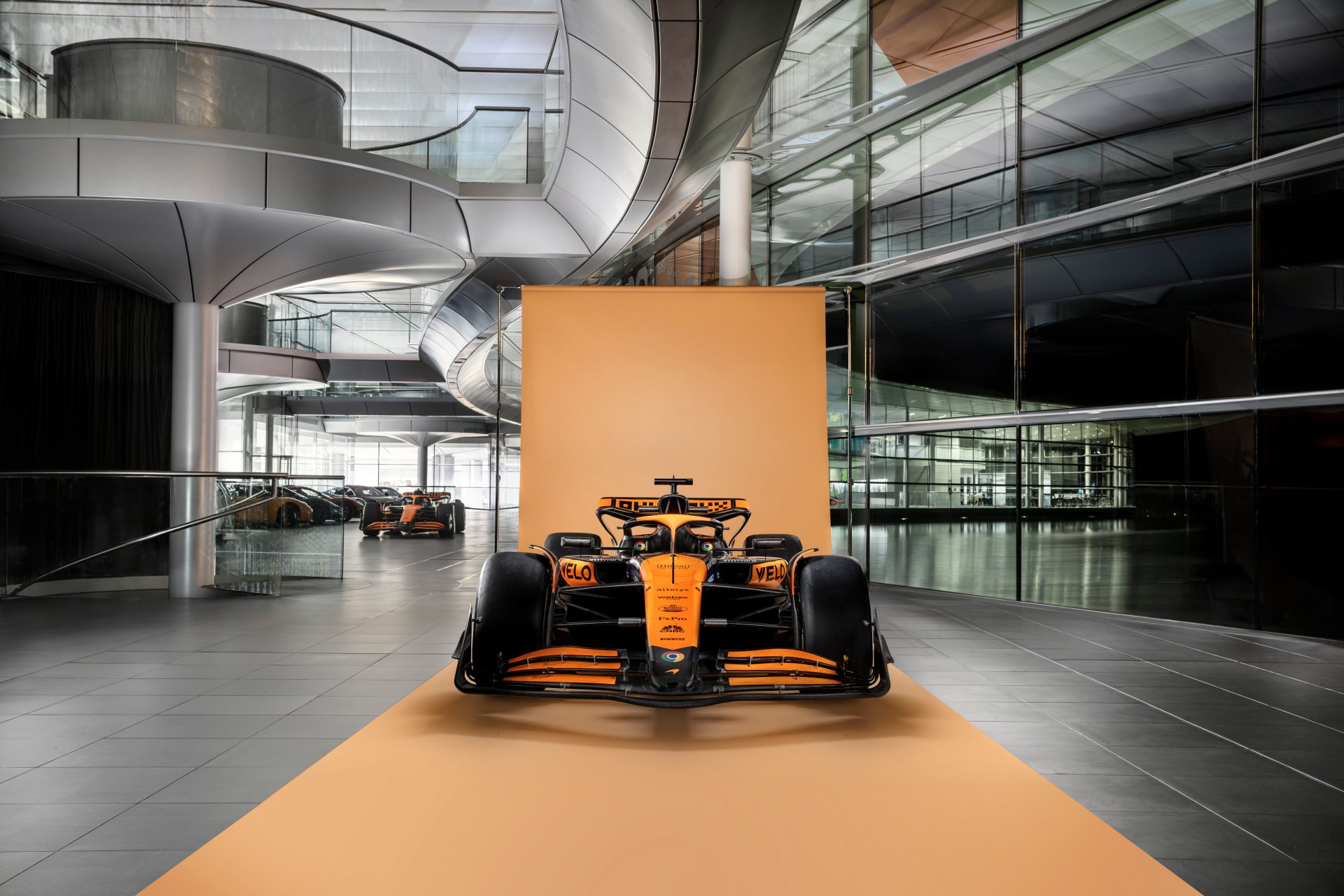 HD desktop wallpaper featuring the McLaren MCL38 Formula 1 car displayed in a modern, architectural venue with reflective surfaces.